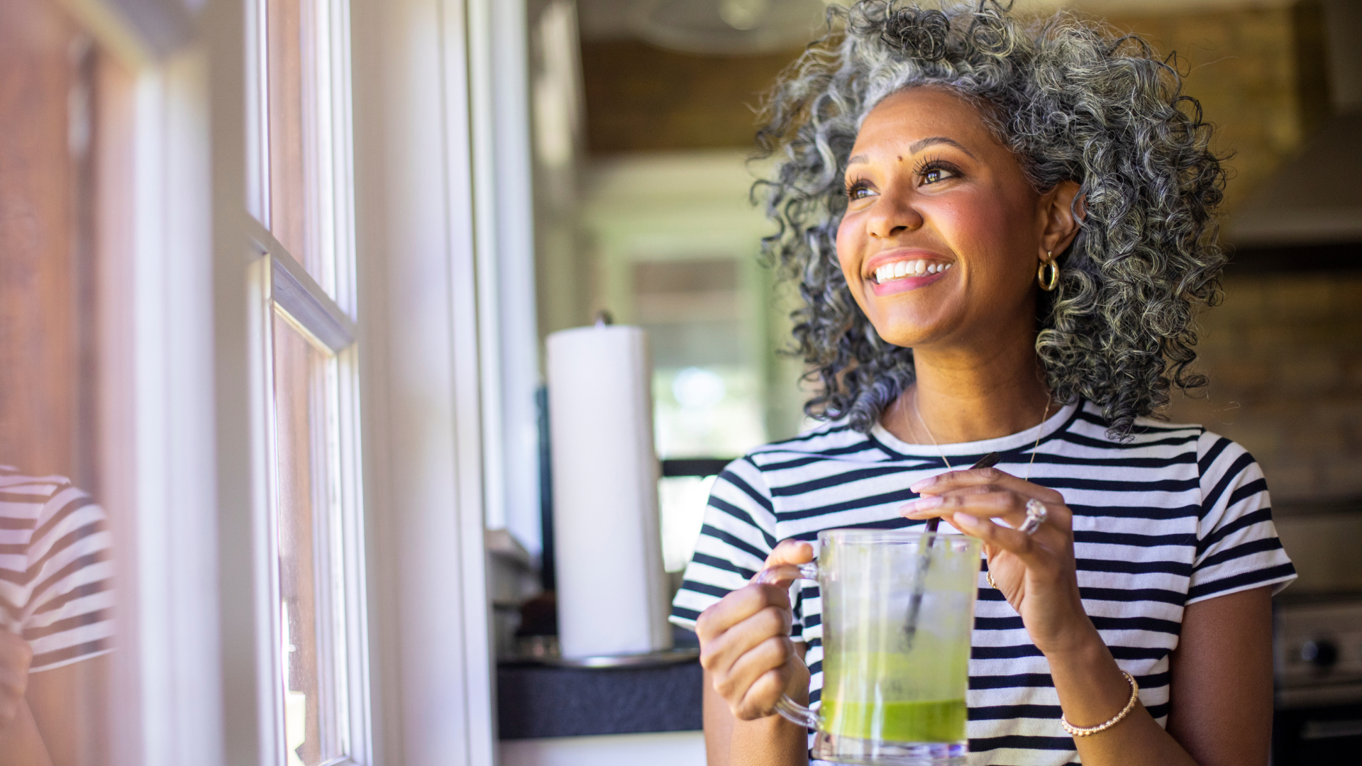 A smiling woman holding a green smoothie beside a sunlit window